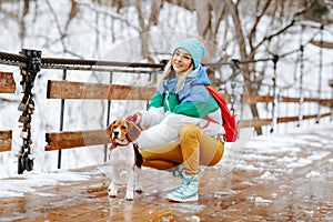 Portrait of cheerful young woman in warm stylish clothes and bagpack posing with her beagle dog on the bridge in winter