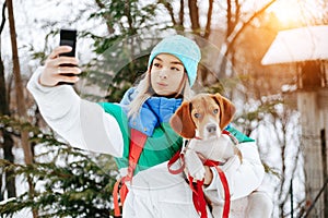 Portrait of cheerful young woman in warm stylish clothes and bagpack take selfie on smartphone with her beagle dog in