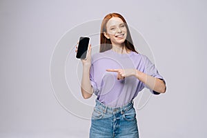 Portrait of cheerful young woman wearing casual clothes pointing with finger on blank mobile screen.