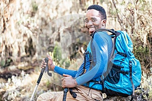 Portrait of a cheerfully smiling African-American Ethnicity young man sitting with a backpack and trekking poles and resting in