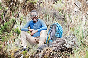 Portrait of a cheerfully smiling African-American Ethnicity young man in sunglasses sitting with a backpack, resting in the forest