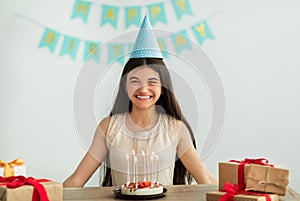 Portrait of cheery Indian teen girl in party hat sitting at table with gift boxes and birthday cake with candles at home