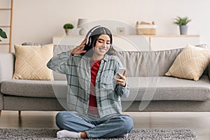 Portrait of cheery young Arab woman in headphones listening to music on mobile device, sitting on floor at home