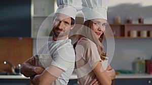 Portrait of chef and assistant in hats standing back to back on kitchen