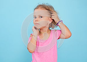 Portrait of cherubic blue-eyed little girl titivating touching short curly fair hair, wearing pink jumpsuit, bracelets.