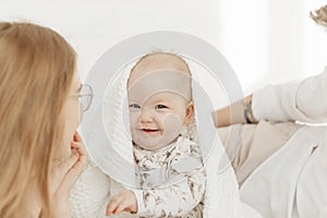 Portrait of cherubic smiling happy blue-eyed plump baby infant toddler covered with white blanket sitting on bed linen.