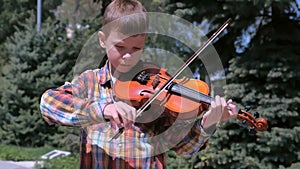 Portrait of child boy is playing the violin standing in park on pine background.