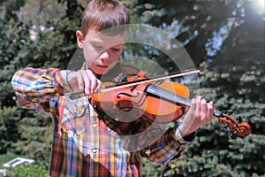 Portrait of child boy is playing the violin standing in park on pine background.