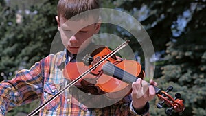 Portrait of child boy is playing the violin standing in park on pine background.