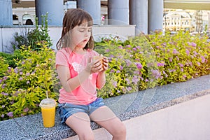 Portrait of a child with a burger and orange juice. Cute girl eating a cheeseburger outdoors. Fast food