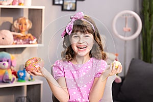 Portrait of child girl choosing between two donuts in home room. Cheerful school girl playing with cakes indoors. Funny