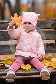 Portrait of a child girl sitting on a bench in autumn city park and playing with maple leaves. Beautiful nature, trees with yellow