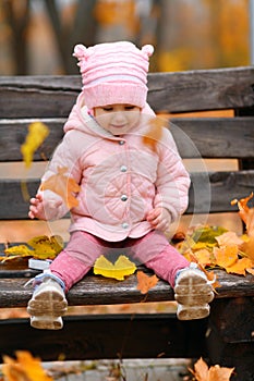 Portrait of a child girl sitting on a bench in autumn city park and playing with maple leaves. Beautiful nature, trees with yellow
