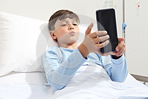 Portrait of child lying alone in bed in hospital room looking at smartphone