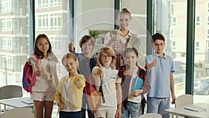 Portrait of the children stand with the teacher in the middle of the class and show a finger up feeling happy and