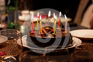 A portrait of a chocolate birthday cake with burning candles on it on a festive dining table. The lit candles all have different