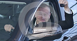 Portrait of chubby Caucasian man bragging car keys at camera and smiling. Happy owner sitting in vehicle and looking out