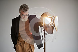 Portrait of classy bearded middle aged man dressed elegantly standing next to studio spotlight while posing for camera