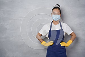 Portrait of a cleaning lady wearing yellow rubber gloves and medical protective face mask keeping arms on hips, looking