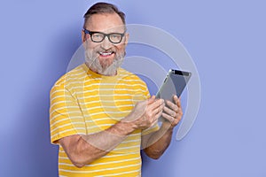 Portrait of clever pensioner persone with beard wear yellow t-shirt in glasses hold tablet read email isolated on violet photo