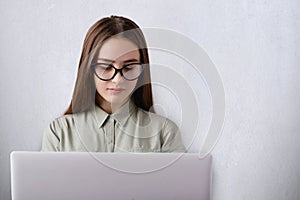 A portrait of clever young girl wearing shirt and glasses being stylish working with her laptop looking down at the screen of the