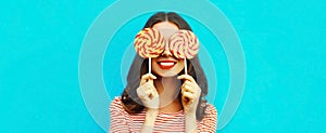 Portrait close up of cheerful smiling young woman covering her eyes with lollipop on blue background