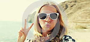 Portrait close up of happy young woman blowing her lips sending sweet air kiss stretching hand for taking selfie on sea background