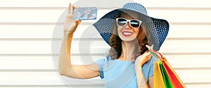 Portrait close up of smiling young woman taking selfie picture by smartphone with shopping bags wearing blue summer straw hat