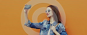 Portrait close up smiling young woman taking a selfie picture by smartphone wearing a denim jacket on a orange background