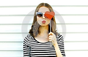 Portrait close-up woman blowing red lips sending sweet air kiss hiding her eye with red heart shaped lollipop on white wall