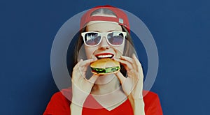 Portrait close up of young woman eating burger wearing baseball cap, sunglasses over blue