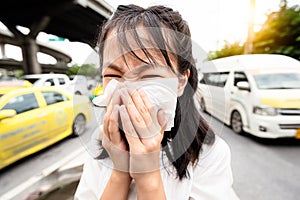 Portrait closeup of child girl blowing nose in paper handkerchief,asian woman sneezing in a tissue in the city street,toxic fumes