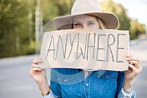 Portrait closeup of woman hopefully waiting passing car in forest holding cardboard poster on roadside. Lady in hat