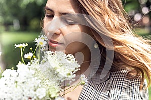 Portrait closeup of young charming woman smelling and enjoying aroma of bouquet plucked white wild flowers. Walking in