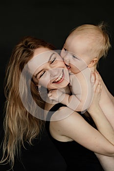 Portrait closeup of young smiling mother holding and embracing cute naked baby on black background. Happy parent