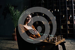 Portrait of cofident young business woman wearing elegant eyeglasses making chess move sitting in armchair in dark