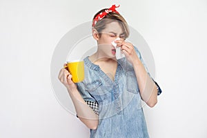 Portrait of cold sick young woman in casual blue denim shirt with red headband standing holding drink and tissue and cleaning her