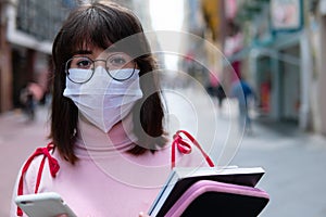 Portrait of college girl walking in the city center. Woman wearing face mask with cellphone in hands and carrying school