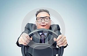 Portrait of a comical businessman in a dark suit and glasses holding a car steering wheel in his hands, on a blue background