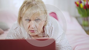 Portrait of concentrated absorbed senior woman surfing Internet on laptop lying in pajamas on bed at home. Relaxed