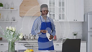 Portrait of concentrated African American man in apron and earphones talking at video chat on laptop holding milk bottle