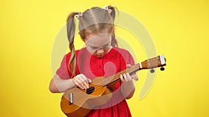 Portrait of concentrated baby girl with pigtails playing ukulele at yellow background. Focused interested Caucasian