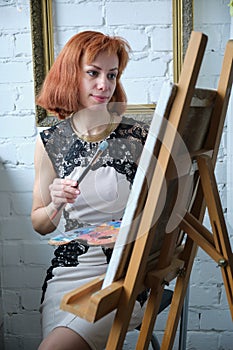 Portrait of concentrated beautiful young adult redhead woman painter holding art palette and painting on canvas with brush at home