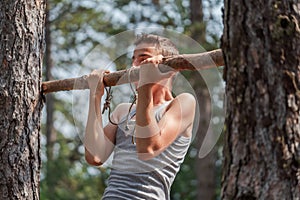 Portrait of concentrated boy in T-shirt doing pull ups on horizontal wood bar outdoors in forest.