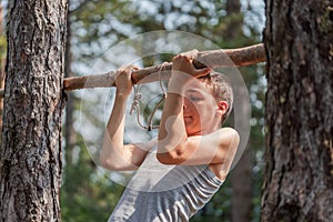 Portrait of concentrated boy in T-shirt doing pull ups on horizontal wood bar outdoors in forest.