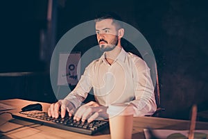 Portrait of concentrated businessman stay at night in dark loft office at evening typing working on computer sit table