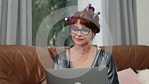 Portrait of concentrated Caucasian senior woman in eyeglasses surfing Internet on laptop sitting on couch at home
