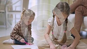 Portrait of concentrated charming Caucasian girl with pigtails painting sitting on floor with blurred baby sister