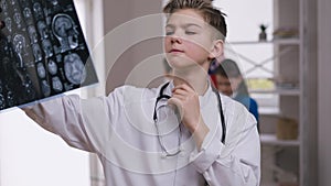 Portrait of concentrated little doctor examining brains X-ray thinking. Teenage Caucasian boy in white medical gown