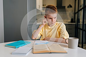 Portrait of concentrated little pupil boy sitting at table, pensive looking away, holding pen, taking notes, doing